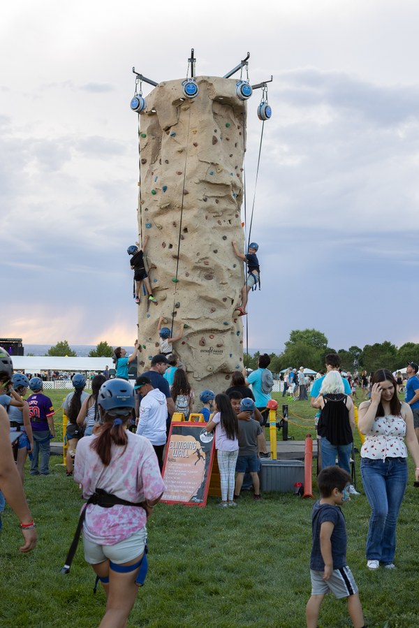 Children climbing a tall outdoor rock-climbing wall at a crowded park event with safety harnesses and helmets.