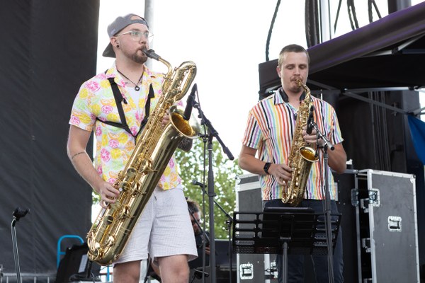 Two male musicians playing saxophones on an outdoor stage during a live performance, one with a baritone sax and the other with an alto sax.