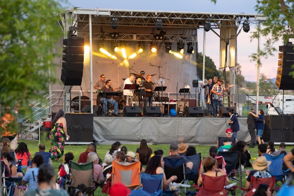 Westside Summerfest Outdoor concert with a band performing on stage as an audience sits on lawn chairs and blankets in a park setting.
