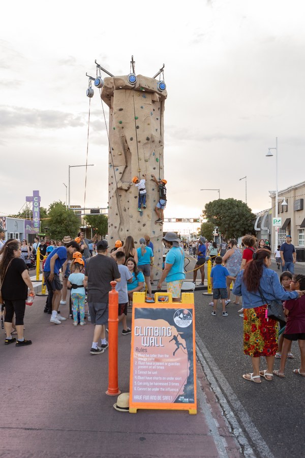 Crowd gathered around a tall outdoor climbing wall with two climbers ascending, set up on a city street.