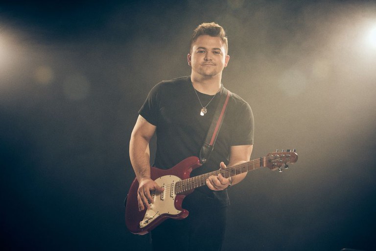 Man holding a guitar standing in front of a black background.