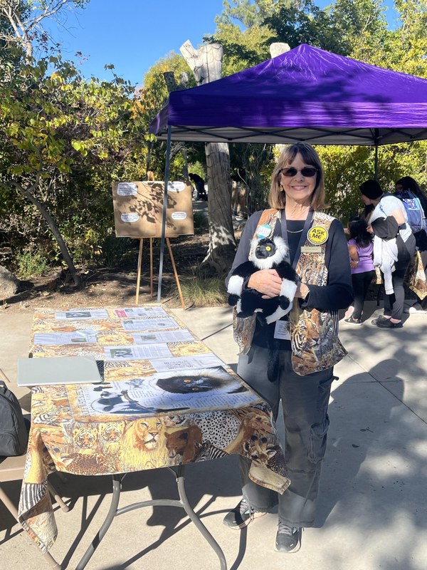 Photo of a woman holding a stuffed animal in front of information booth. On the table are a variety of photos, papers, and infographics.