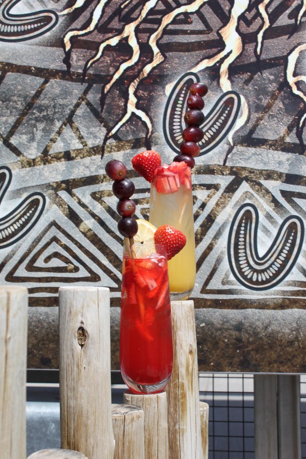 Photo of two classes of mimosas resting on fence posts in front of an elaborate mural. One is pink and strawberry flavored, one is yellow and pineapple flavored. Each has a skewer with grapes sticking out of the top.