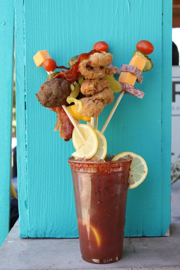 Photo of loaded bloody mary drink in front of a blue painted fence. The drink is loaded with skewers of toppings including lemons, a meatball, cheese, etc.