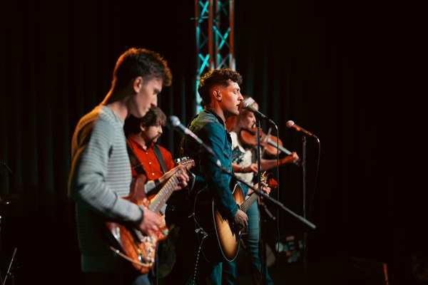 Four musicians performing on stage with guitars and microphones under warm lighting and a dark backdrop.
