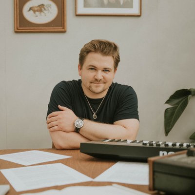 Photo of man in black short sleeve t-shirt sitting at a wood desk, smiling at the camera. There are papers scattered on the desk and a electric keyboard on the side.