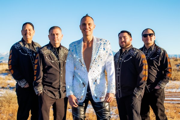 Five men standing outdoors in a desert landscape, with one wearing a sparkling white jacket and the others in matching black embroidered shirts.