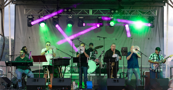 Seven-piece band performing live on an outdoor stage with colorful green and purple lights illuminating the scene.