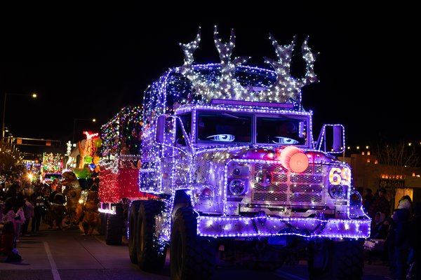 Military truck decorated with purple and white Christmas lights, featuring illuminated reindeer antlers and a red nose, driving in a nighttime holiday parade.