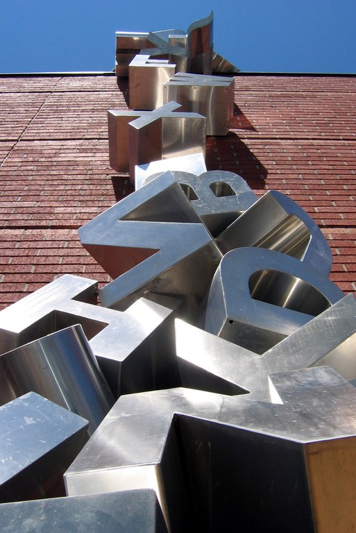 Three-dimensional stainless steel capital letters of the English alphabet falling from the side of the library and piling up on the ground. 