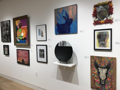 Multiple artworks hanging on white walls in an art gallery, including paintings, collage, and a glass work.