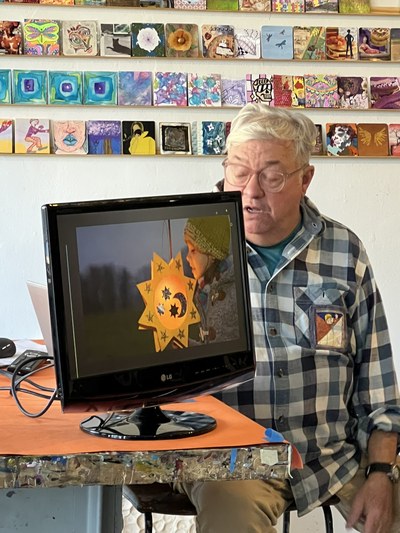 Artist Robert M. Allen seated behind a monitor displaying an image of a lantern, sharing the history of lanterns.