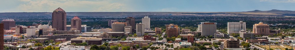 A grand skyline view of Downtown Albuquerque taken from the east facing the west. A line of tall buildings with the green Bosque and expansive mesa appear in the background. It's a bright, sunny day with a few clouds in the far distance.