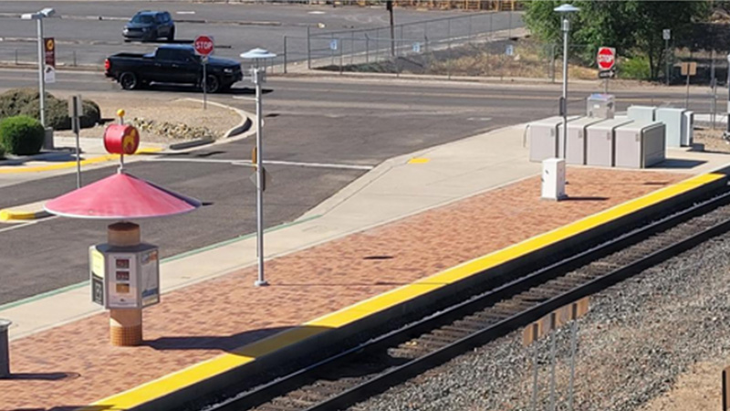A distance view of a section of train track and the loading platform with a covered info kiosk and a row of gray metal lockers. A mostly empty parking lot can be seen in the upper left.