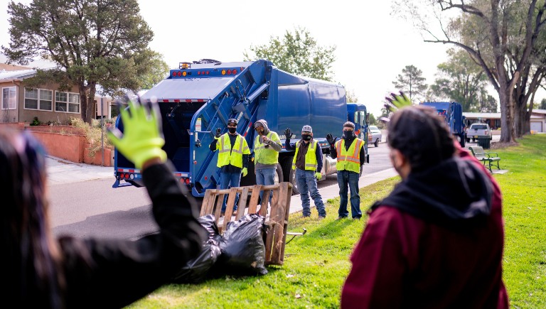A family in the foreground waves a team of Solid Waste employees standing in front of a blue trash truck.