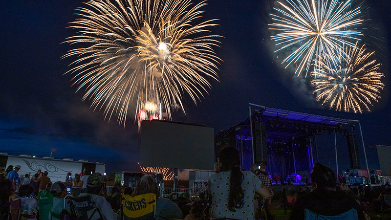 A group of people seated in lawn chairs in front of a stage at night gaze up as fireworks explode in the sky.