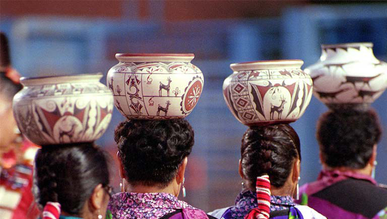 The backs of women with traditional hairstyles holding traditional pottery above their head.