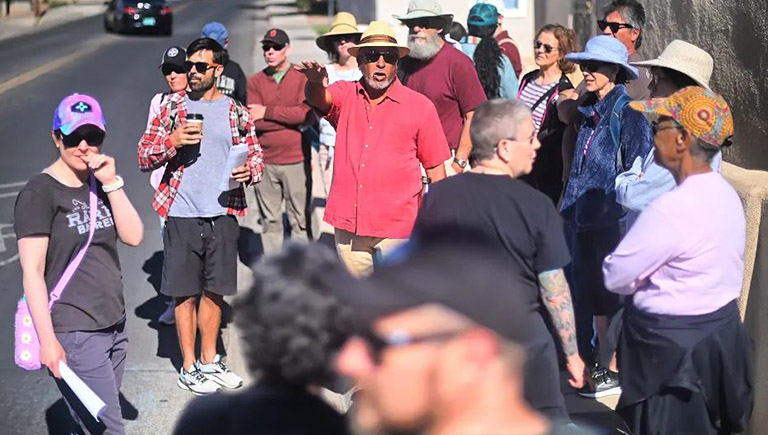 A group of people stand on the corner of a street and look off-camera. It's a bright, hot day. Most people are wearing shorts, hats, and sunglasses.