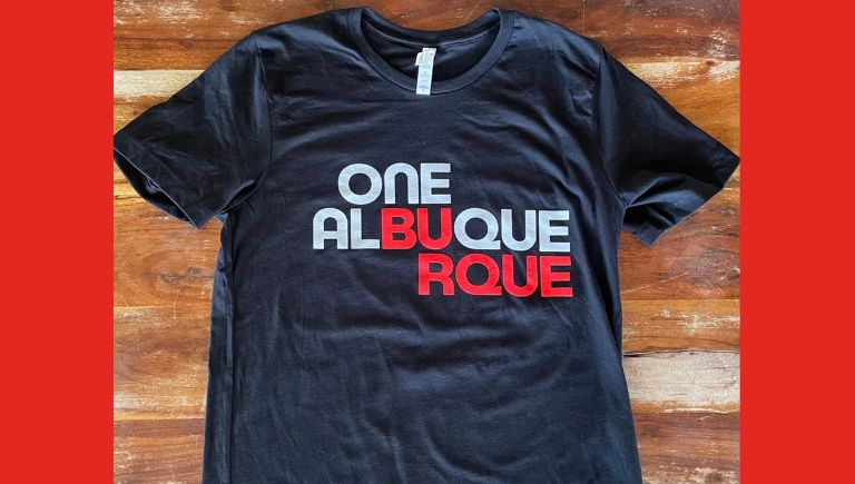A black t shirt on a wood plank background with the red and white One Albuquerque logo.