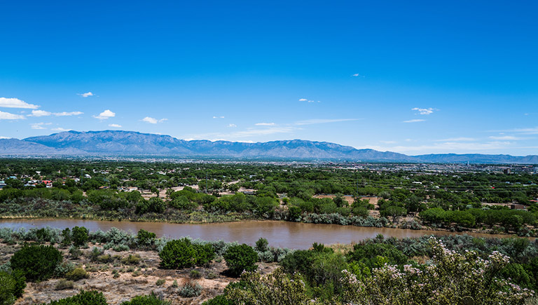 West Central Albuquerque New Mexico with the Rio Grande. The bosque and Rio Grande are in the foreground and the Sandia Mountains are visible in the background. A few homes peek out through the trees.