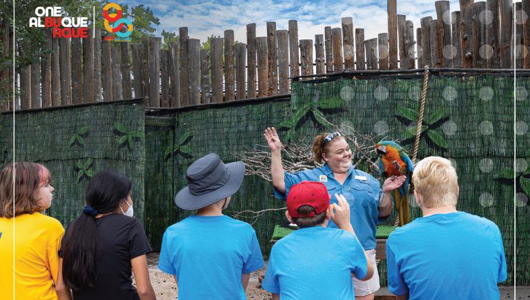A woman in a uniform polo holds a scarlet macaw in front of 5 sitting children.