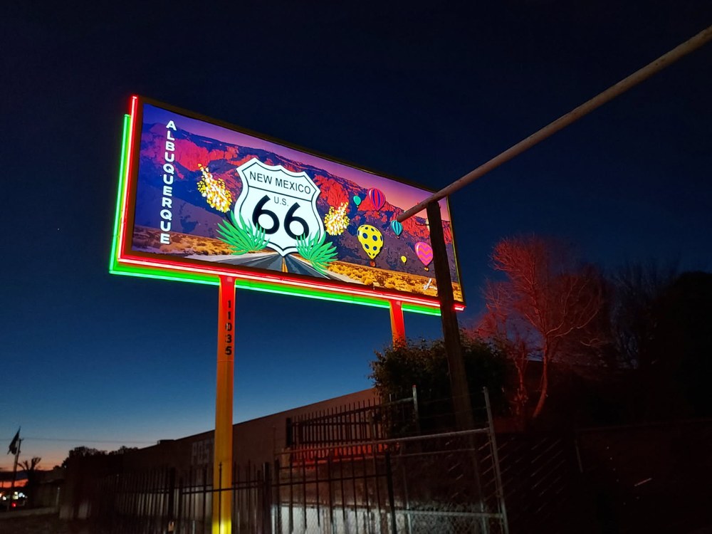 Night time photo displays a neon billboard at 11035 Central Avenue NE, with a giant vintage U.S. Route 66 badge emblem, the sunset-hued Sandia Mountain range as a backdrop, with recognizable Albuquerque icons of colorful hot air balloons and blooming yucca.  
