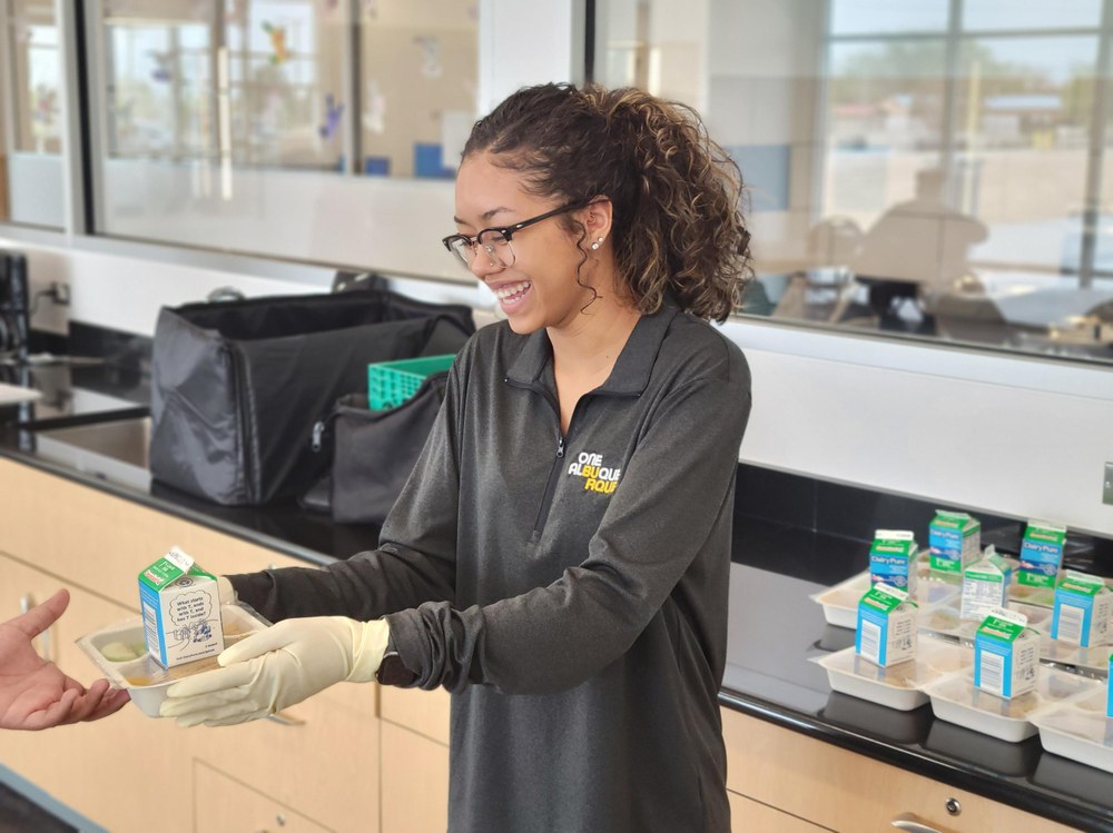 A city employee hands a packaged lunch to a child in a park.
