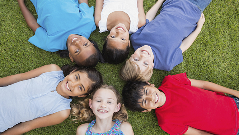 A group of children laying on the grass arranged in a circle with their heads in the center, all looking up and smiling at the audience.