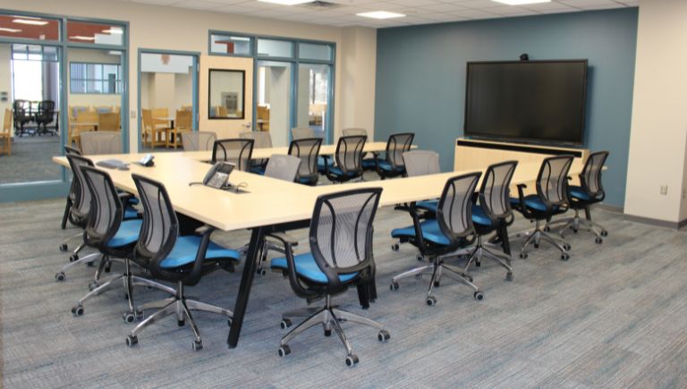 A U shaped conference room table surrounded by blue chairs in front of a large TV.