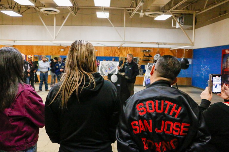 A man with a black jacket that says "south San Jose Boxing" stands in the foreground of the Jack Candelaria boxing gym. In the background, Mayor Keller is speaking at a podium about the coming renovations.