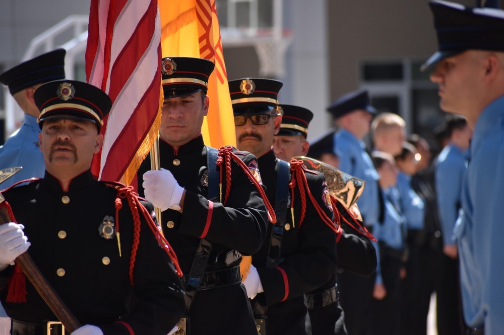 Four firefighters in dress uniforms carrying the USA flag, the NM state flag, and axes.