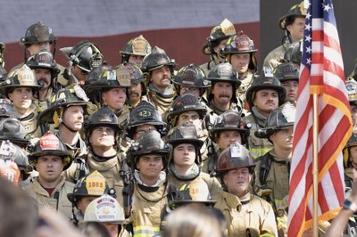 2026 911 Memorial Stair Climb Firefighters A group of firefighters in full gear with photos of fallen firefighters on their hemets standing behind the U.S. flag.