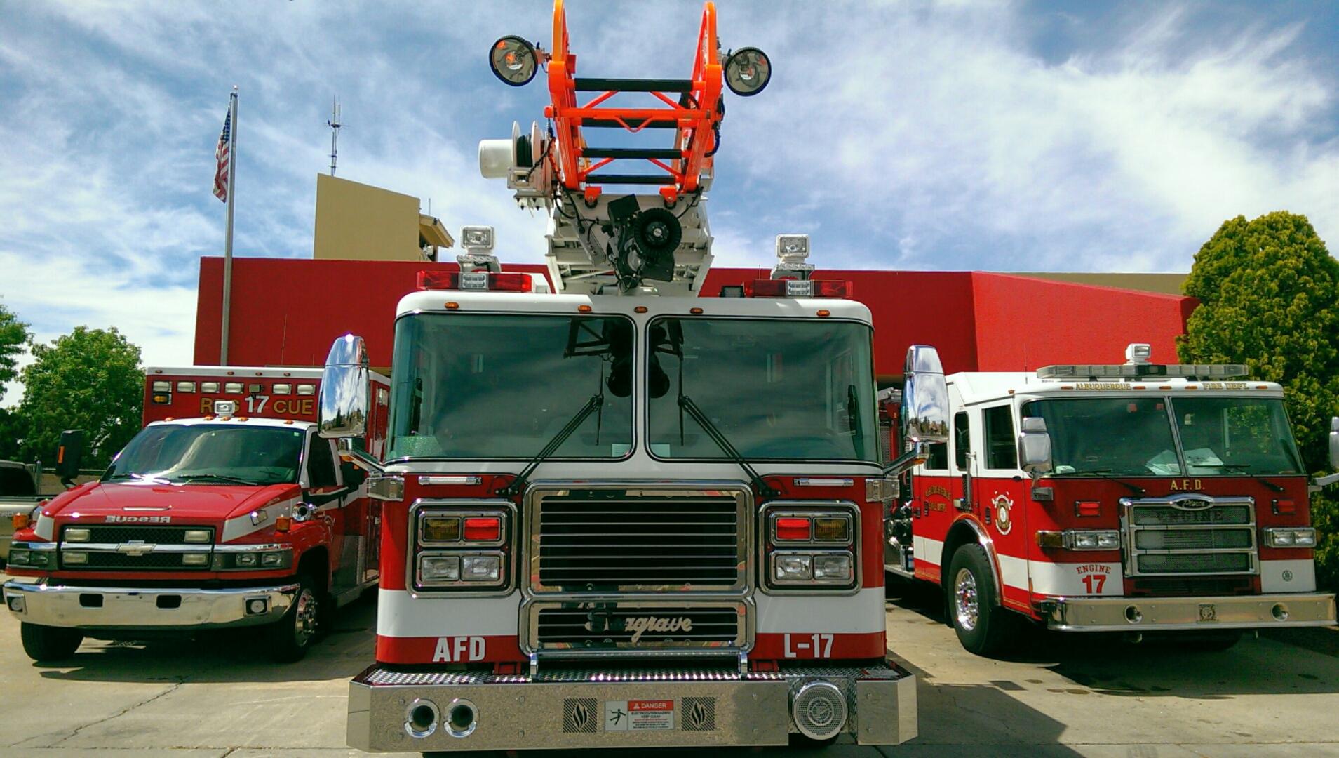 Three Albuquerque Fire Rescue vehicles (two fire trucks and an EMT vehicle) outside of an AFR building, all facing the audience and lined up in a row with the middle vehicle closer than the flanking two.