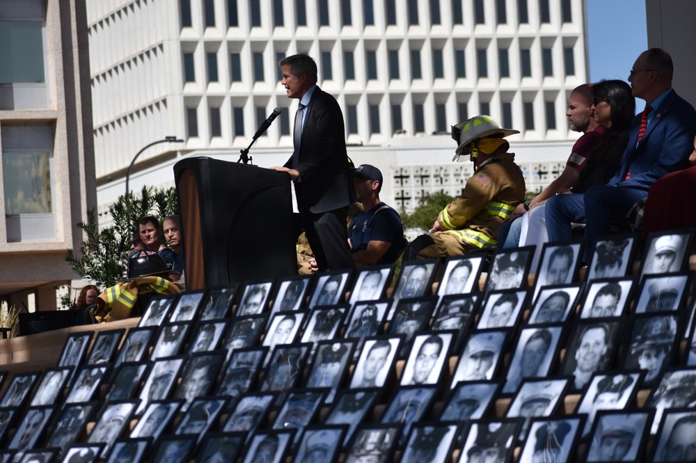 Photos of fallen firefighters lined up facing the crowd with Mayor Keller speaking from a podium behind them.