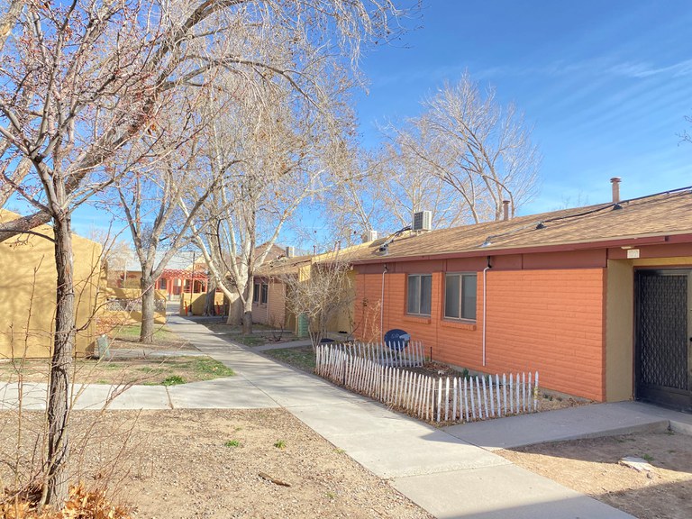 A colorful row of yellow and orange single-story brick apartments with a sidewalk and trees in between.