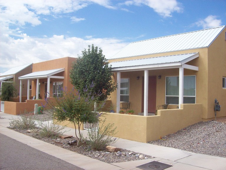 Two homes on a street, one with copper colored stucco and one with tan stucco. Front porches face the street and one house has a tree in the front yard. A lavender bush is in the foreground along the street