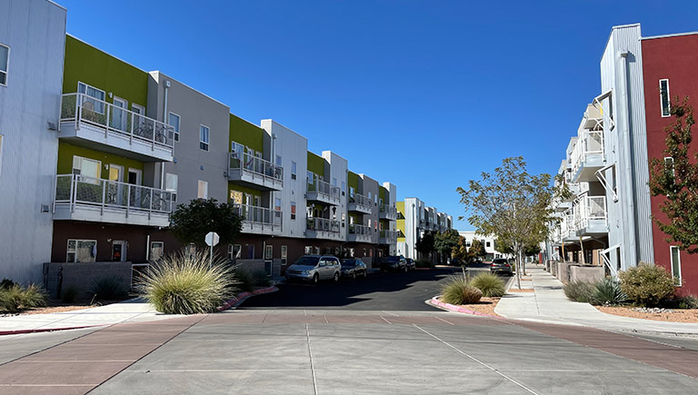 An exterior view down a street featuring a three-level apartment complex painted green, brown, and red. Cars are parked along the side of the access street. The sidewalk in front of the units is lined with trees.