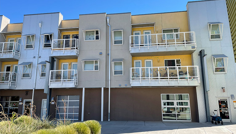 A street view of a three-level apartment complex, painted in brown and yellow. The units are modern with metal accents and have balconies on each unit.