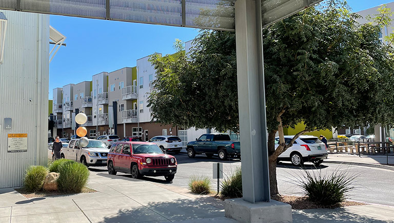 A view of a three-level apartment complex taken from the sidewalk under a covered patio area near the units. A woman in a dark dress walks along the sidewalk, passing multiple vehicles parked along the side of the row in front of the complex. Trees and shrubs line the sidewalks.