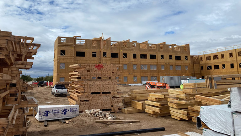 An apartment complex in the middle of construction. The complex has been framed and the general structure is complete. Stacks of construction wood and other building materials are organized in the foreground.