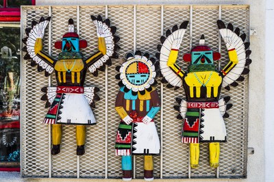 Three brightly colored Native American wood carvings of people dressed in traditional Native American clothing and feathers, 2 with bird heads and 1 with a round head surrounded by feathers.