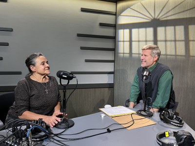 Mayor Tim Keller and an older woman, the guest on the podcast, engaged in conversation on a recent episode of the One ABQ & You Podcast. Both individuals are seated at a triangular gray table and have microphones in front of them.
