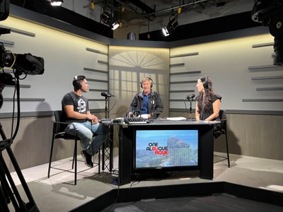 Mayor Tim Keller, seen center, and Leah Black, seen right, and a podcast guest, seen left, sit at a triangular black table. Mayor Keller is speaking and the other two individuals are listening. All are wearing black headphones and have microphones in front of them. Small portions of the studio camera equipment can be seen in the edges of the frame on the left and right.