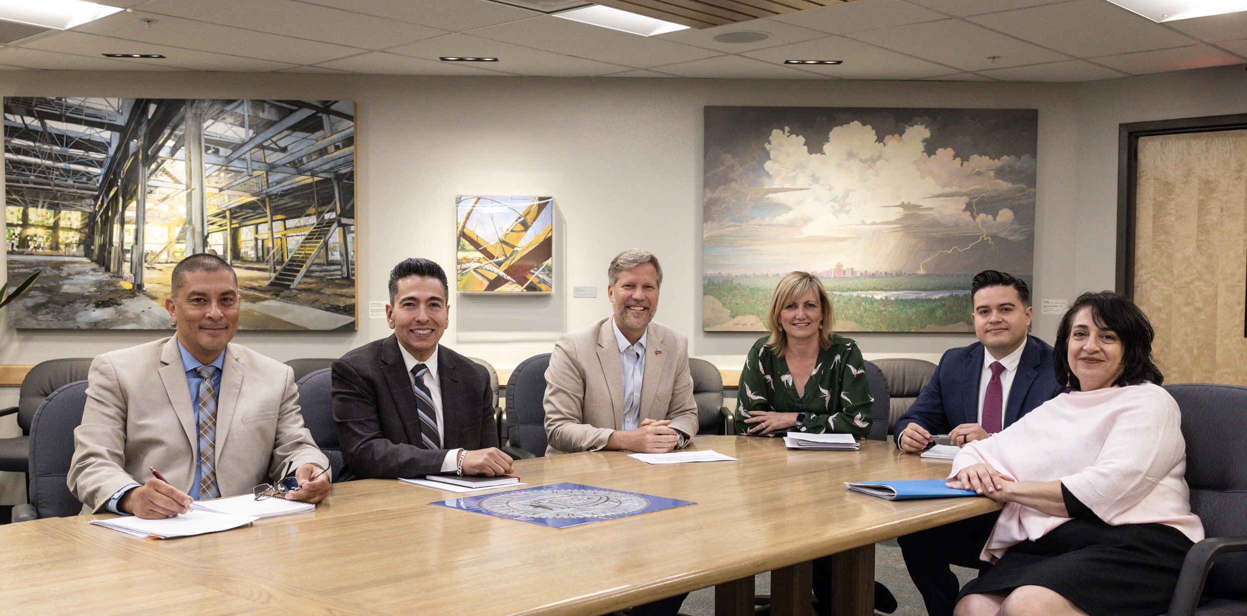 The Mayor's Executive Team gathered around a large conference table. All are seated and smiling at the audience.