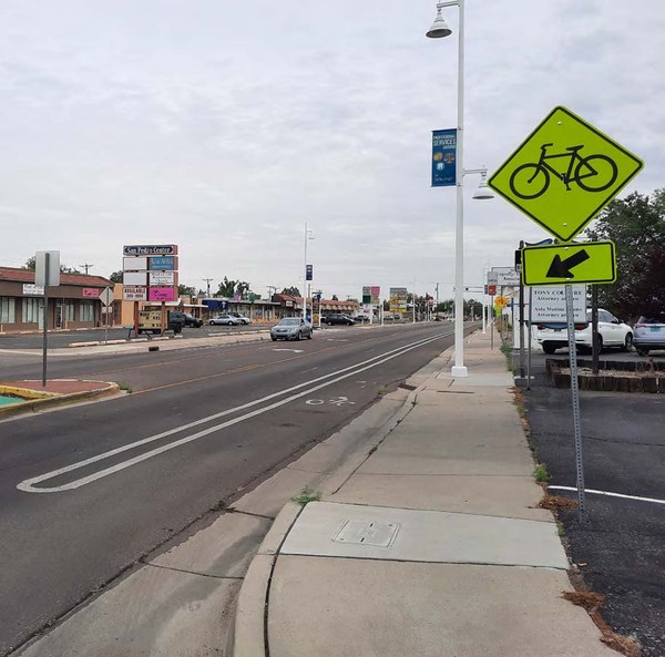 There is a bike lane with a striped buffer between the bike lane and the vehicle driving lane. There is also a bright yellow diamond-shaped sign with a bicycle symbol and an arrow pointing toward the bike lane. The sign is adjacent to the sidewalk.