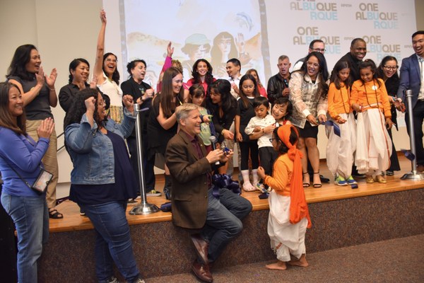 A group of people including Mayor Keller, Councelor Nichole Rogers, and Carlo James Aragón on a stage in front of a screen with the OEI One Albuquerque logo.