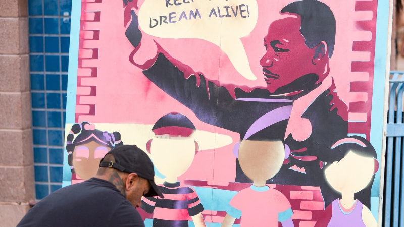 A man painting a mural of Martin Luthor King Jr. saying "I have a dream" with 3 children from different races stand in front holding hands.