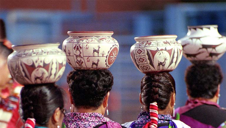 A Photo of the backs of women with traditional hairsyles holding traditional pottery above their head.