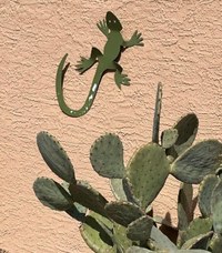 A metal lizard sculpture mounted to a light tan adobe wall. A cactus plant appears in the foreground.