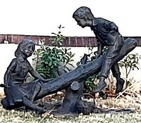 A bronze, realistic metal artwork sculpture of two children playing on a teeter totter.